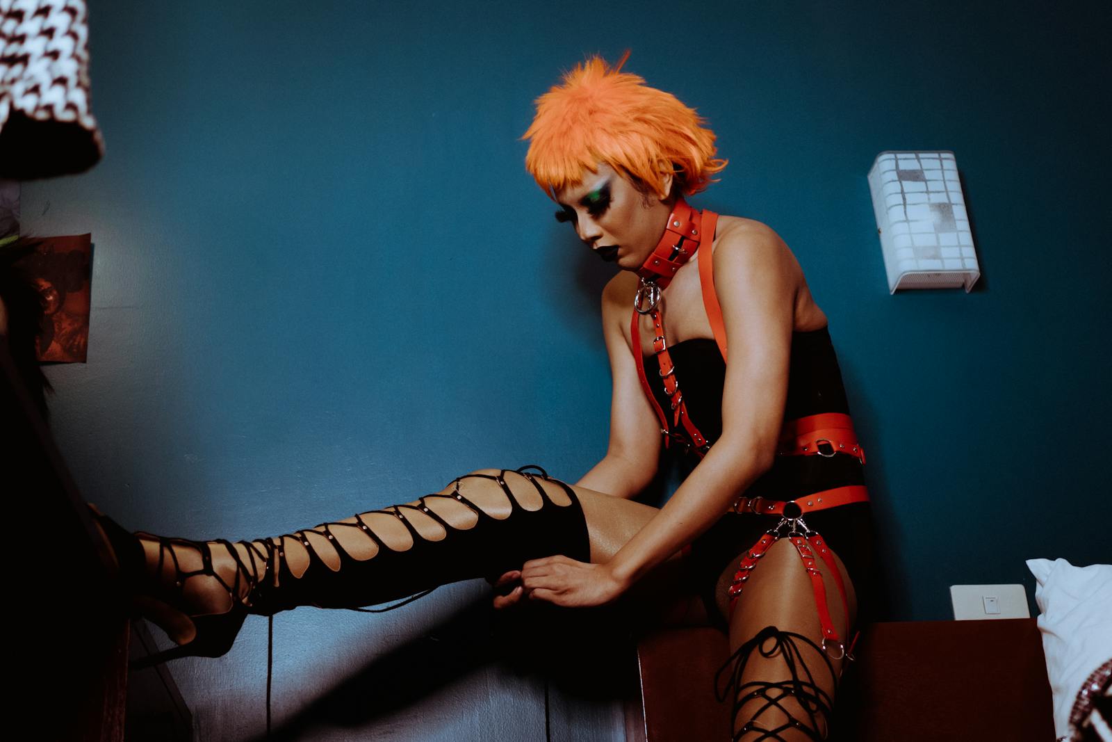 Extravagant female with dark makeup and orange wig wearing black bodysuit and leather harness raising leg on vanity table and zipping thigh high shoe in dark bedroom